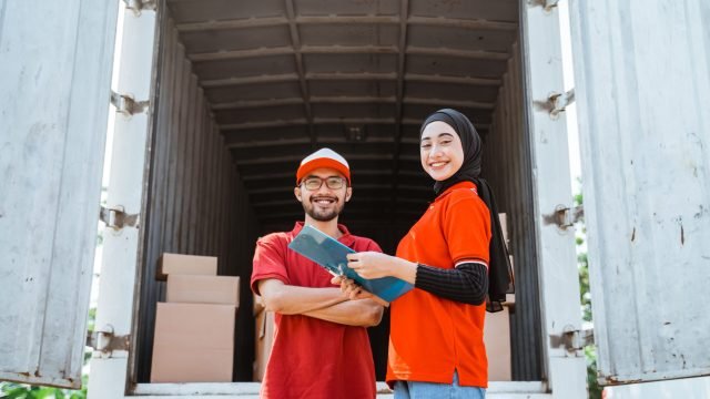 Smiling female worker standing holding clipboard with delivery man on background of cardboard packages inside container truck