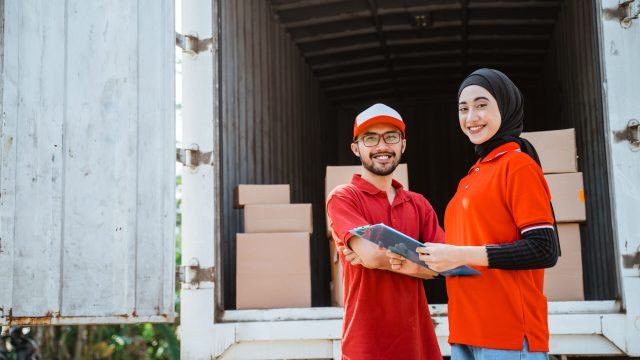 delivery man and smiling asian woman workers standing against background of cardboard packages inside container truck