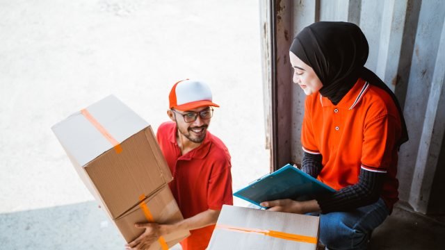 male workers lifting boxes and women workers in headscarves with clipboards checking in containers